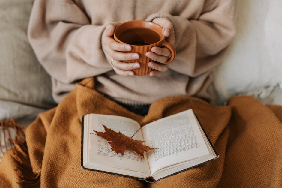 pexels-photo-9969147-9969147 A woman enjoys a cozy autumn day, holding a tea mug over an open book with a maple leaf.